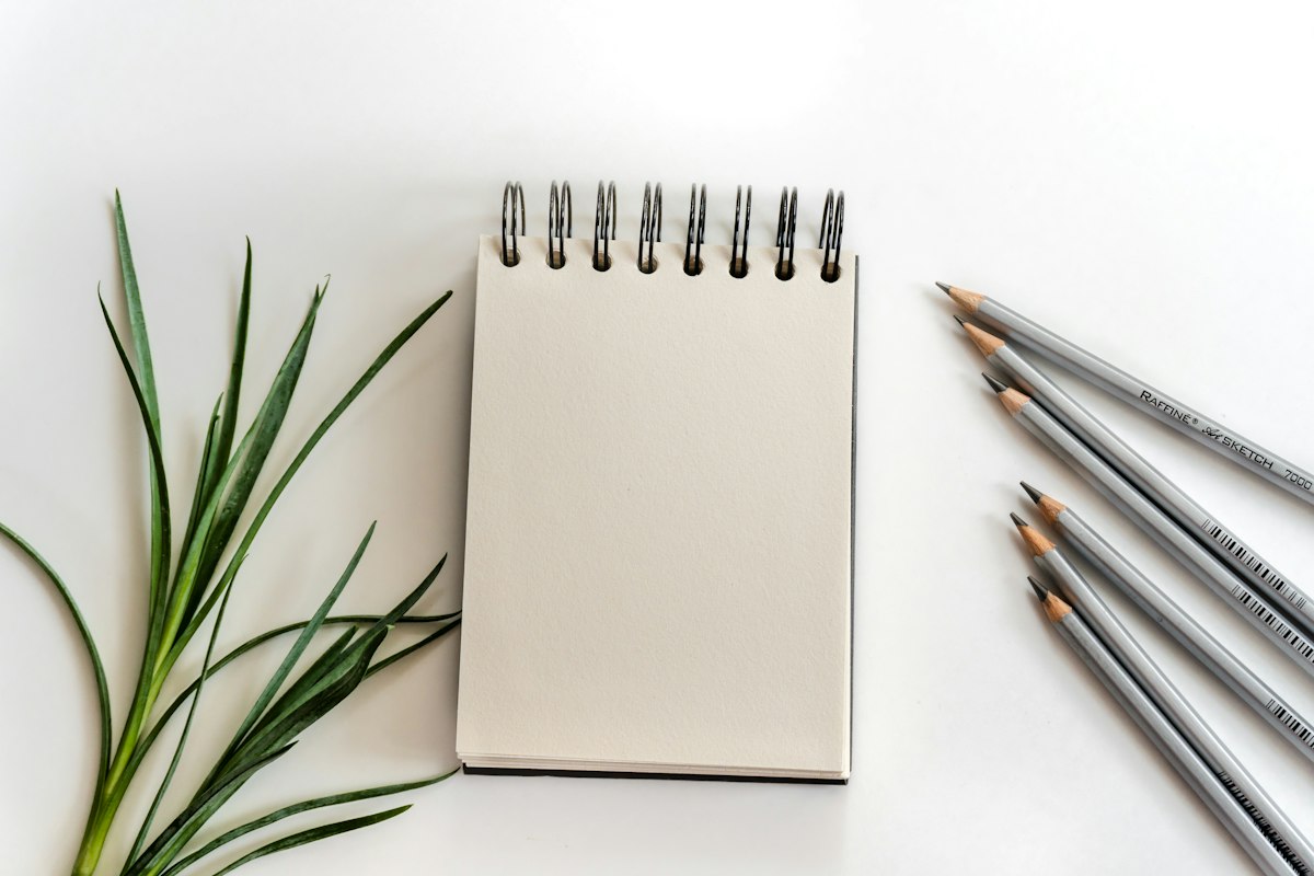 Caderno em branco, folhas verdes e lápis sobre uma superfície branca; imagem ilustrativa